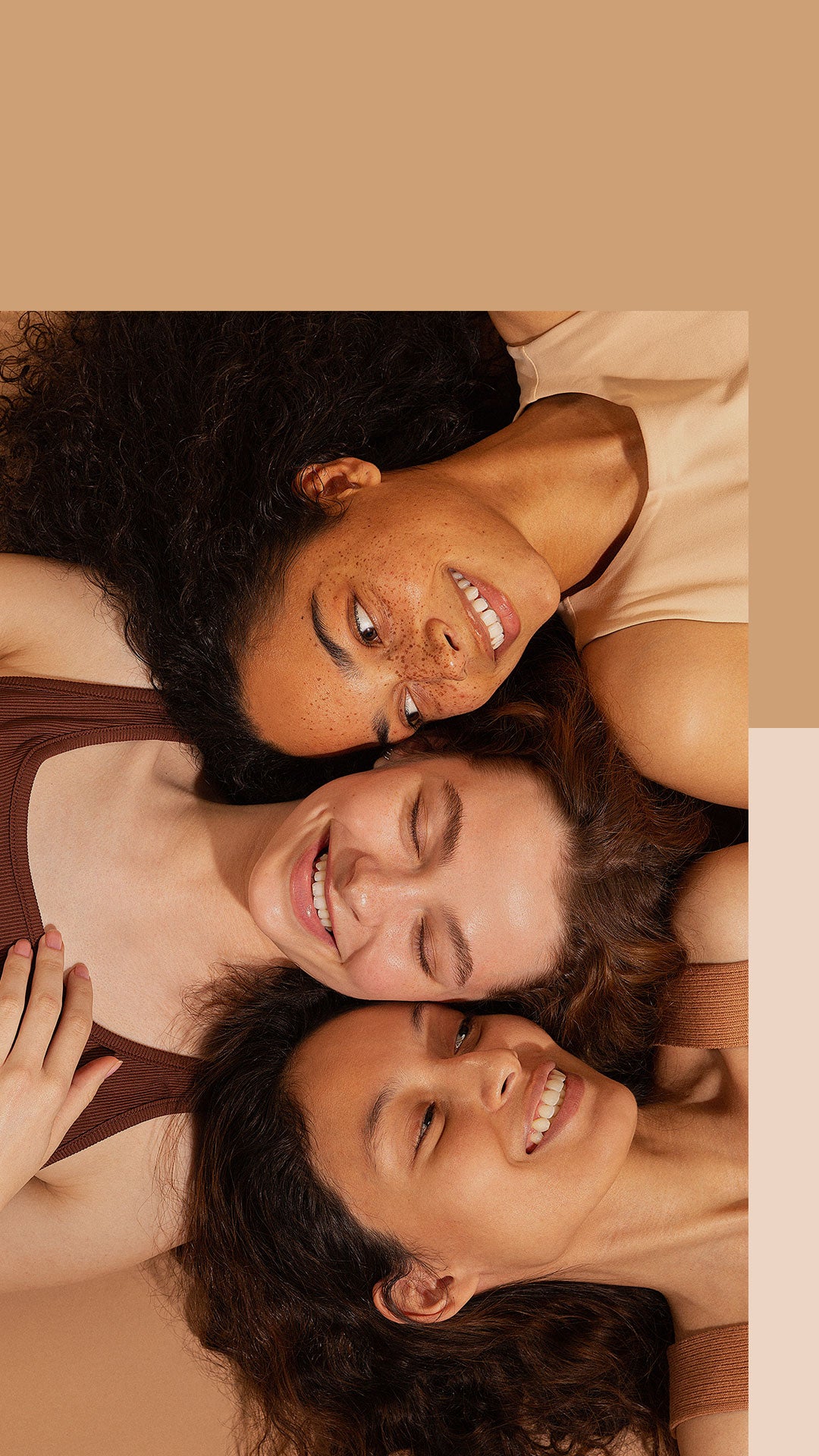 Close-up headshot of 3 young women lying on their backs smiling at eachother against a neutral background